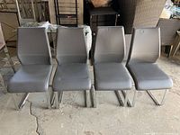 Front view of four grey faux leather dining chairs with chrome cantilever legs. Shown side by side on a concrete floor in a storage area.