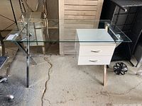 Full side view of the glass top desk showing clear glass surface, chrome X-frame legs, and white drawer unit.