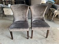 Front view of two gray upholstered dining chairs with slightly flared backs and wooden legs showing minor scuffs.