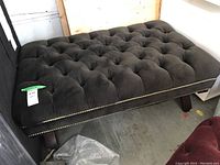 Full view of rectangular tufted ottoman on four dark wooden legs, showing top and side with metal stud edging.