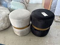 Two round ottomans side by side, one beige microfiber and one black microfiber, both with gold metal bands around the center, placed on concrete floor in warehouse setting.