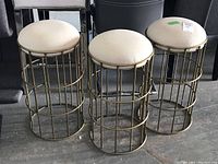 Three metal stools with gold-tone vertical bar bases and round off-white upholstered seats grouped together on floor