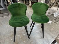 Front view of two green velveteen upholstered stools with black metal legs and a footrest bar, highlighting shape, material, and condition.