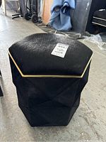 Black hexagonal storage ottoman covered in animal hide style material, showing the angled sides and gold trim on lid.
