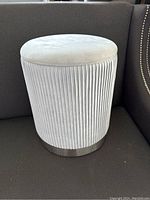 Full side view of light gray round microfiber storage ottoman with pleated vertical lines and metallic base trim