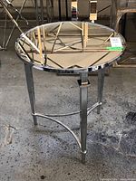 Photo showing the circular side table with a reflective mirrored glass top and a shiny metallic frame on three legs with a tripod-like base.