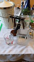 Photo of the bar set items including glass bowl with tongs, shot glass with card suits, ceramic ice bucket, and bar tool stand with tools on a table covered with white cloth.