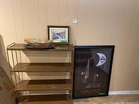 Front view of the metal frame wooden bookshelf with four shelves. On the top shelf, a decorative bowl with autumn leaf pattern and a framed picture are visible. To the right, framed Mardi Gras poster is partially visible but not part of lot.