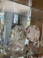 Photo showing two china platters on stands and four glass items including two wine decanters and two glass vases inside a cabinet.