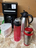 Braun drip coffee machine with glass carafe, stainless steel thermos, red plastic tumbler, stainless steel tumbler, and set of coffee filters on countertop in front of pegboard.