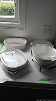 Four Corningware dishes and two metal stands displayed on countertop, three have glass lids.