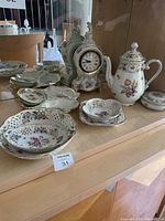 Overall view of all porcelain serving pieces and clock arranged on shelf.