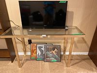 Console table with clear glass top and polished metal frame, shown with a TV and three Frank Sinatra albums below it. Tape label on table top showing dimensions 48 x 15 x 26 inches.