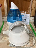 Photo showing white electric kettle with handle and cord in front and a blue and white humidifier with translucent tank in background, placed on a surface with manual behind them.