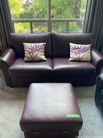 Leather burgundy couch with two cushions and matching ottoman in front of window.