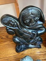 Frontal view of black soapstone carving depicting a mother holding her child, showing detailed facial features and clothing textures, with a visible crack on the stone.