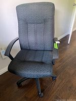 Front-facing view of the black fabric office chair showing the cushioned backrest, seat, and plastic armrests on a hardwood floor.