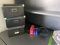 Three stacked black storage boxes with silver metal corner protectors and label holders alongside a black tape holder, two boxes of envelopes, and a multi-outlet extension cord.
