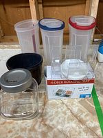 Photo of three plastic cereal dispensers, glass jar, black container, and boxed rotating card tray laid on a floor.