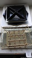 Multiple serving platters stacked including black divided ceramic platters on top of white platters and a gold-tone wire basket tray placed in front.