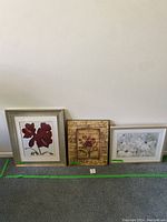 Three pieces of artwork displayed side by side on the floor against a wall: two framed floral prints and one decorative solid piece in the center. The left framed print features a red flower, the middle piece has vintage Parisian floral design, and the right framed print shows white flowers.