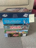 Stack of six boxed games: Pavilion glass chess, Killer Bunnies Blue Starter Deck, Risk, Monopoly Animal Crossing, Guelph on Board, visible from side view on carpeted floor.