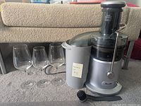 Wide view of Breville Juice Fountain Plus juicer with attached pulp container, three clear Schott Zwiesel wine glasses, and part of the cleaning brush on carpeted floor.