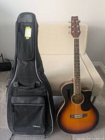 Front view of Fender acoustic guitar and black soft case standing on floor against couch.