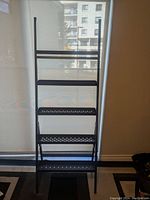 Full front view of the Ikea black steel ladder shelf standing against window blind showing all four perforated metal steps and top bar.