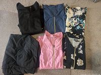 Five Jaanuu scrub tops folded on carpet floor showing variety of colors and styles including solid black, dark blue with zipper, pink with zipper, floral prints.