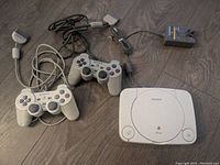 Sony PlayStation One console with two wired controllers and power adapter all shown on floor