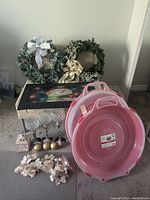 Photo showing 2 green wreaths propped against the wall, pink floral decor pieces in front of a pink large plastic tub, small gold spherical candles, and some candle holders on a table.