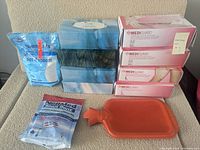 Top view showing 4 boxes of disposable gloves (2 medium, 2 large), 4 boxes of facial tissues stacked, NeilMed sinus rinse packets, orange hot water bottle, and blue bag of Epsom salts.