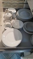 Drawer with stacked white ceramic dishes including plates, bowls, cup, sugar, and cream containers all with a raised bead rim design.