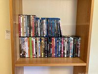 Wooden shelving unit with DVDs and Blu-rays arranged in two rows showing various titles including Mash, Mad Max, West Side Story.