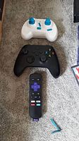 Three controllers on carpet: white game controller with blue thumbsticks, black Xbox controller, and Roku remote with purple directional pad and branded shortcut buttons