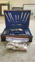 Wooden canteen open showing two compartments of flatware pieces with blue velvet lining and extras outside in a plastic bag.