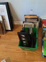 Assorted frames in and around a green bin on wood floor, including black, silver, and gold tones