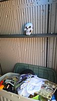 White plastic skull mask on shelf behind storage bin containing Halloween items.