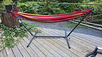 Full view of portable hammock on a black metal stand placed on wooden deck outside, surrounded by greenery.