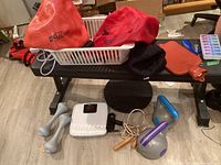 Overview of exercise bench with multiple fitness accessories: red inflatable exercise balls, weighted gloves, jump rope, pumps, dumbbells, and a scale on and near the bench.