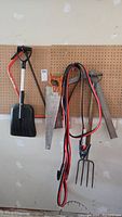 Full view of all items hanging on pegboard including snow shovel, potato fork, two handsaws, two L squares, and jumper cables.