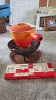 Vintage Le Creuset enameled fondue pot on black metal stand with burner base, showing vibrant orange to red gradient and spouted handle