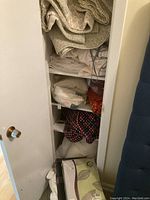 White shelving unit containing various folded linens such as bedding, blankets, pillows, and curtains, in neutral colors and a red and black plaid fabric
