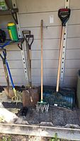 Full view of garden tools against wall including shovel, potato fork, claw Gold cultivator, edger, thatcher, and green snow shovel