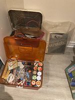 Photo of open amber plastic sewing box showing spools of thread, pins, measuring tape, and scissors inside compartments.
