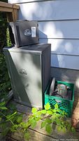 Full view of smoker cabinet on deck next to house siding with feeder unit on top and green crate beside it