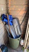 Photo of green garbage can holding blue and gray tarps, wooden-handled edger tool, and hockey stick, with white sprayer bottle beside it.