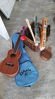 Lot overview showing the Kohala wooden ukulele with blue soft case, three flute recorders, orange and black boxed harmonica, and brown wicker basket on carpeted floor.