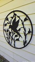 Full view of circular metal wall art featuring hummingbird and floral design hung on beige siding.